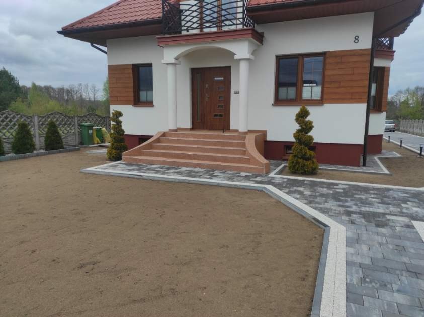 Wykończona posesja przed domem jednorodzinnym z ułożoną kostką brukową, schodami wejściowymi z jasnego kamienia i przygotowanym terenem pod trawnik.