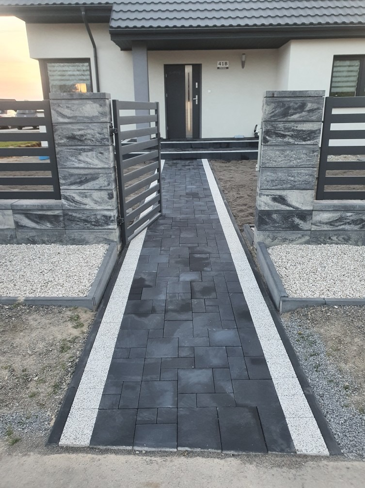 Nowoczesne wejście do domu z ciemnej kostki brukowej, obrzeża z jasnego grysu, otwarte metalowe ogrodzenie z betonowymi słupkami.