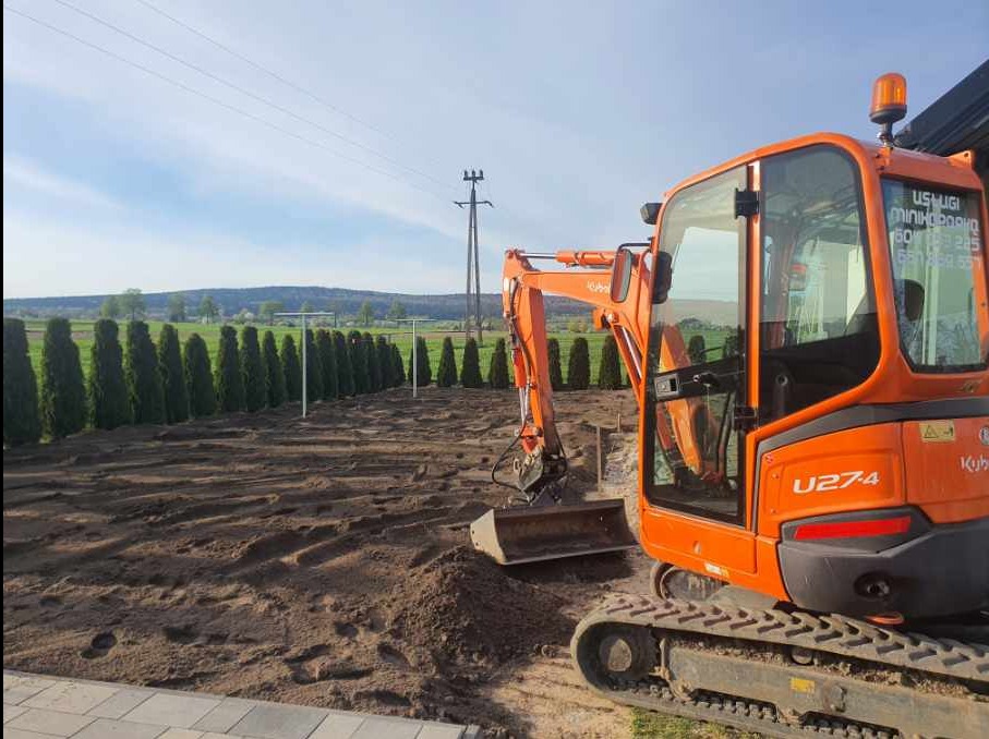 Pomarańczowa mini koparka Kubota U27-4 na nierównym terenie z widocznymi śladami pracy, w tle rząd tuj i słup energetyczny pod błękitnym niebem.