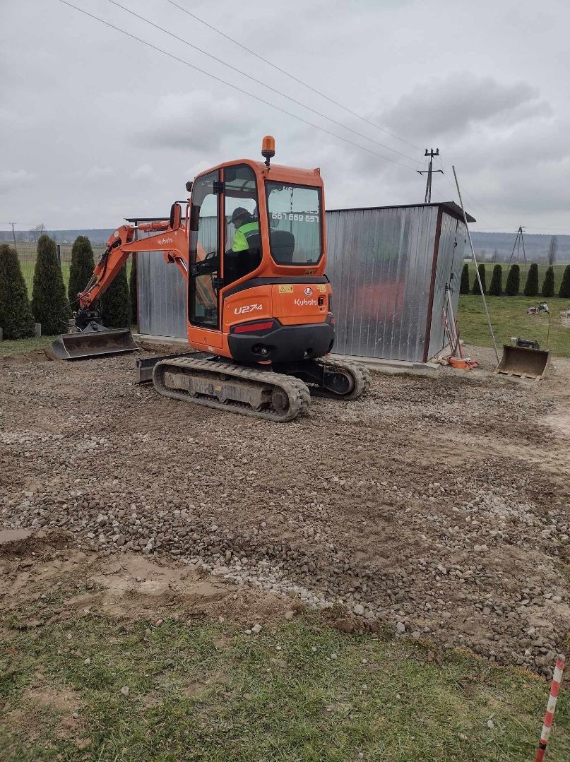 Pomarańczowa mini koparka Kubota U274 z operatorem w kamizelce odblaskowej na żwirowym podłożu, w tle blaszany garaż i tuje.
