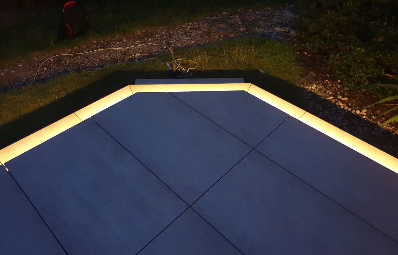 Betonowy taras podświetlony taśmą LED wzdłuż krawędzi, widoczne kable zasilające na trawniku.