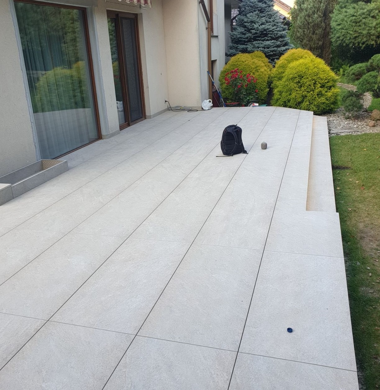 Jasnoszary taras wyłożony dużymi płytami, z widocznym plecakiem i kamieniem na powierzchni, przylegający do domu z zielonym ogrodem w tle.