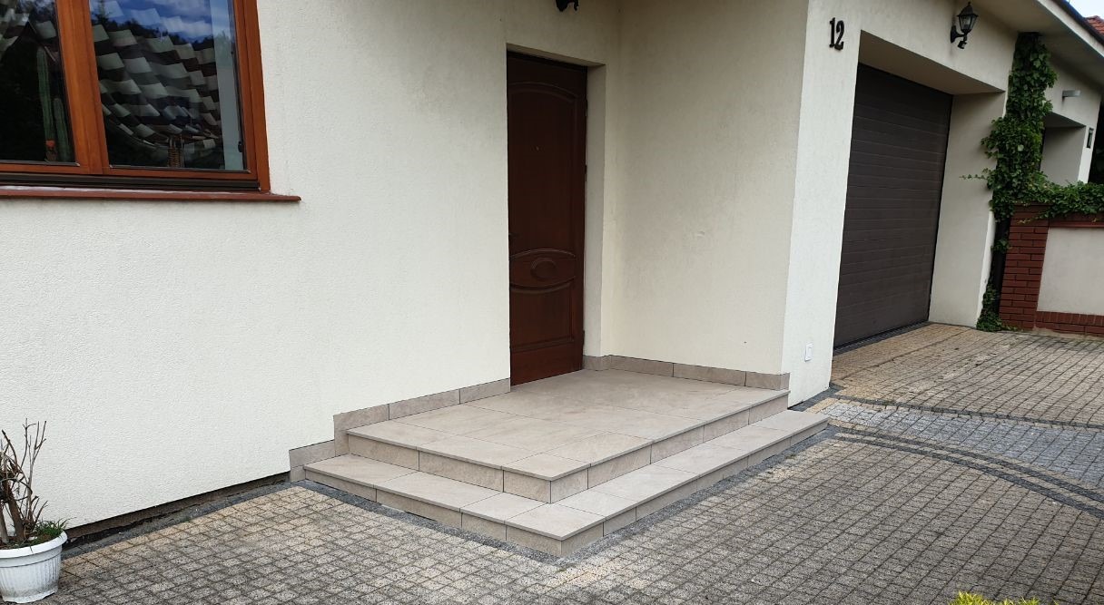 Wyłożone jasnymi płytkami schody wejściowe do domu z brązowymi drzwiami, obok garaż z numerem 12 i pnąca się roślina.