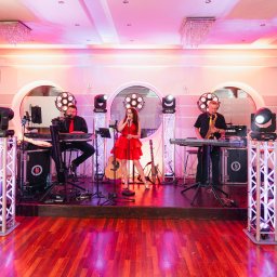Hocus-Pocus.pl - Wesela i Eventy - Zespół muzyczny na scenie: wokalistka w czerwonej sukience, klawiszowiec z brodą i saksofonista, oświetlenie sceniczne, gitary w tle.