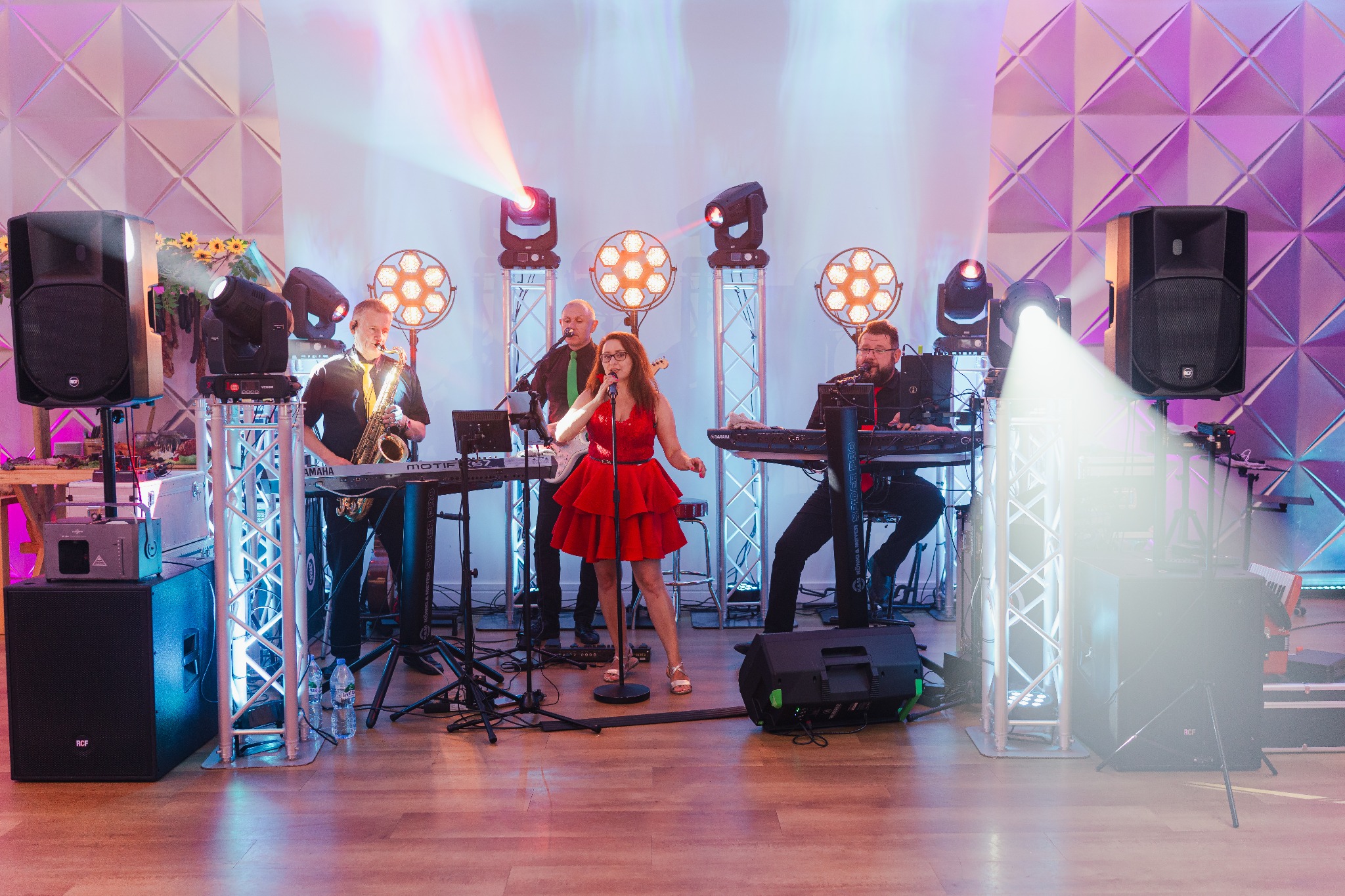 Zespół muzyczny na scenie: wokalistka w czerwonej sukience, saksofonista, gitarzysta i klawiszowiec, oświetlenie sceniczne, białe dekoracje ścienne.