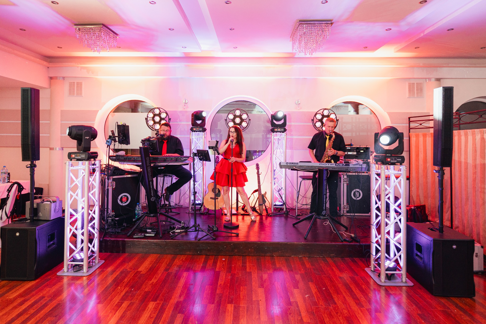Zespół muzyczny na scenie: wokalistka w czerwonej sukience, klawiszowiec z brodą i saksofonista, oświetlenie sceniczne, gitary w tle.