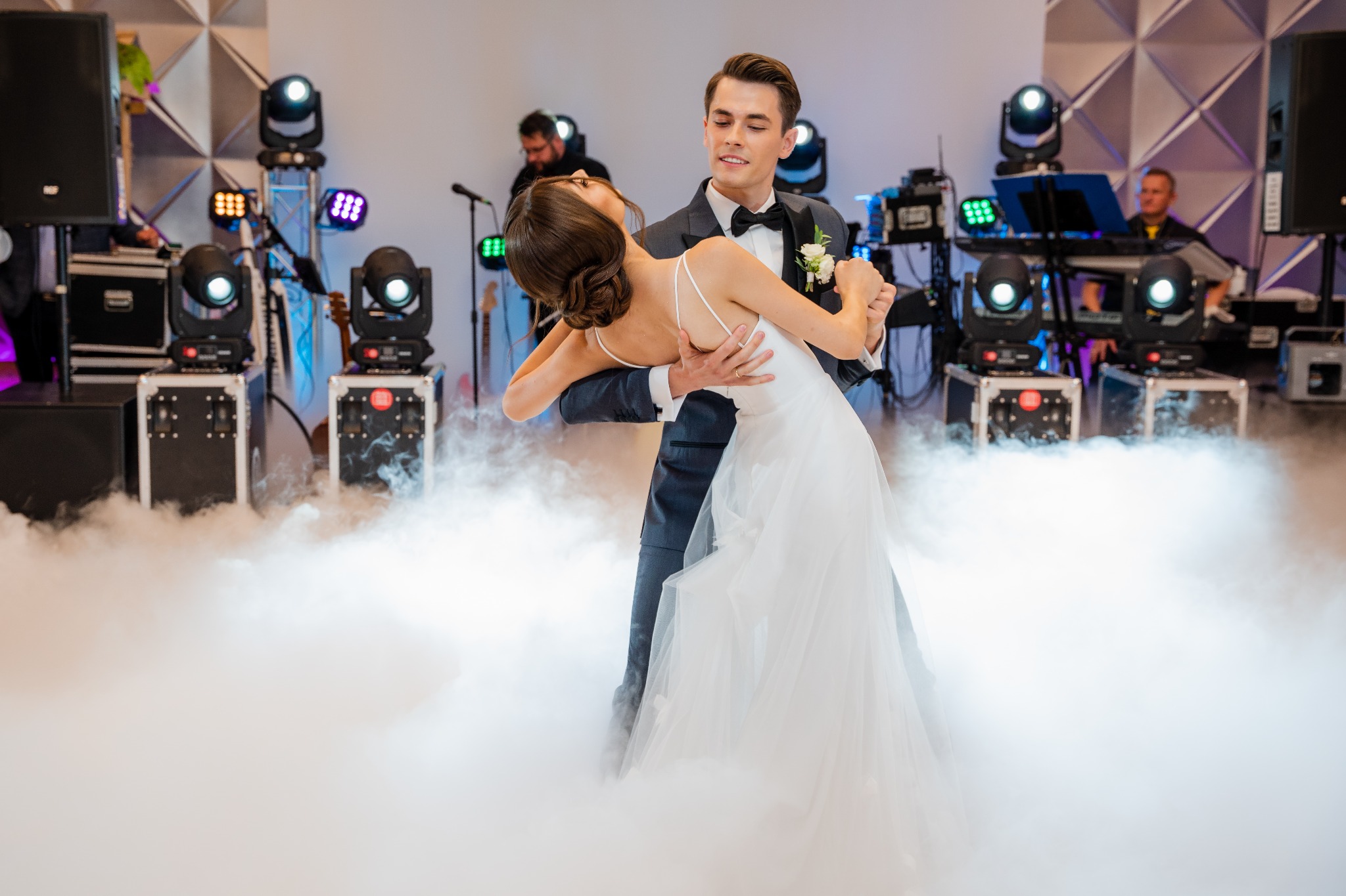 Para młoda tańczy pierwszy taniec w chmurze dymu na tle sceny z oświetleniem i zespołem muzycznym.