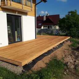 TARAS-MEB Michał Pawlik - Nowo wybudowany taras z jasnego drewna przylegający do białego budynku z czarnymi oknami, widok na ogród i dom w tle.