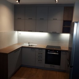 TARAS-MEB Michał Pawlik - Nowoczesna kuchnia z szarymi szafkami, drewnianym blatem, oświetleniem LED pod szafkami wiszącymi i metalową lampą wiszącą, widziana z niskiego kąta.