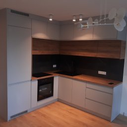 TARAS-MEB Michał Pawlik - Nowoczesna kuchnia w kształcie litery L z szarymi frontami i drewnianymi blatami, czarny panel nad blatem, oświetlenie punktowe i designerski żyrandol, widok z niskiego kąta.