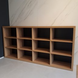 TARAS-MEB Michał Pawlik - Półka drewniana z dwunastoma przegródkami na tle ściany w kolorze ciemnego granatu i ściany wyłożonej płytkami imitującymi marmur.