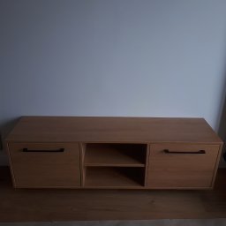 TARAS-MEB Michał Pawlik - Drewniana szafka RTV z dwoma szafkami i otwartą półką pośrodku, z czarnymi, długimi uchwytami, wykonana na wymiar w Krakowie, na tle białej ściany i drewnopodobnej podłogi.