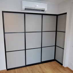TARAS-MEB Michał Pawlik - Nowoczesna szafa przesuwna z czarnymi ramami i matowymi panelami, zamontowana pod klimatyzatorem w jasnym wnętrzu.
