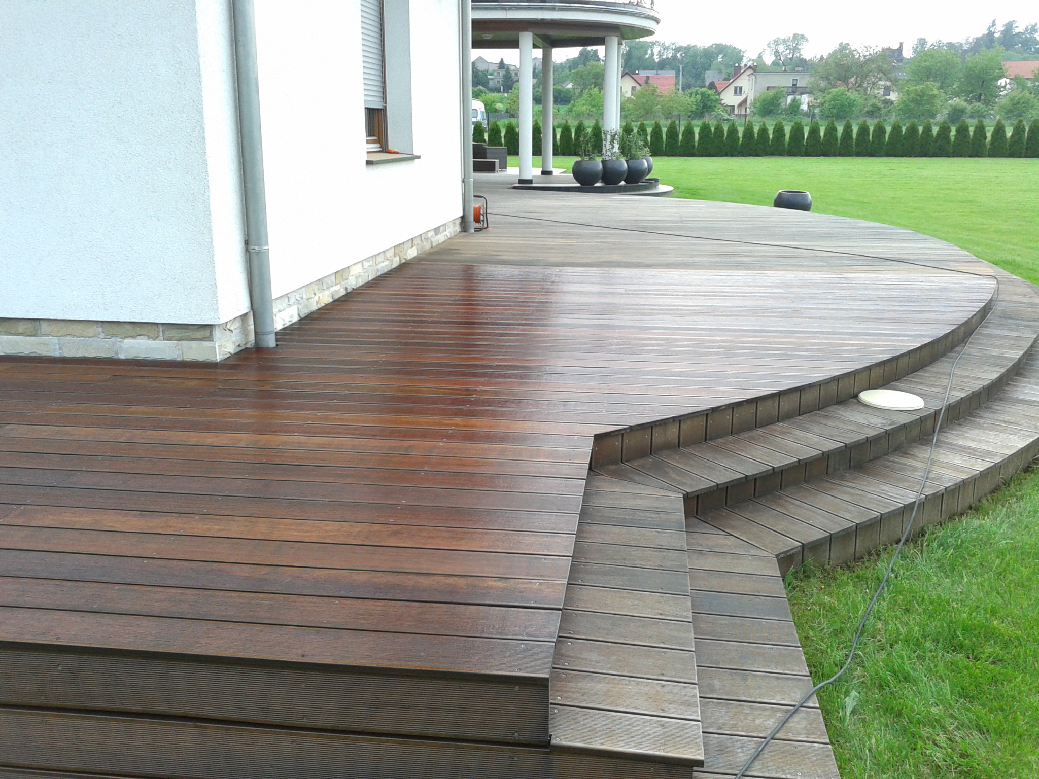 Rozległy taras z drewna egzotycznego z półokrągłymi schodami prowadzącymi do ogrodu. Widoczna ściana budynku z oknem, z tyłu kolumnada z roślinami w donicach.