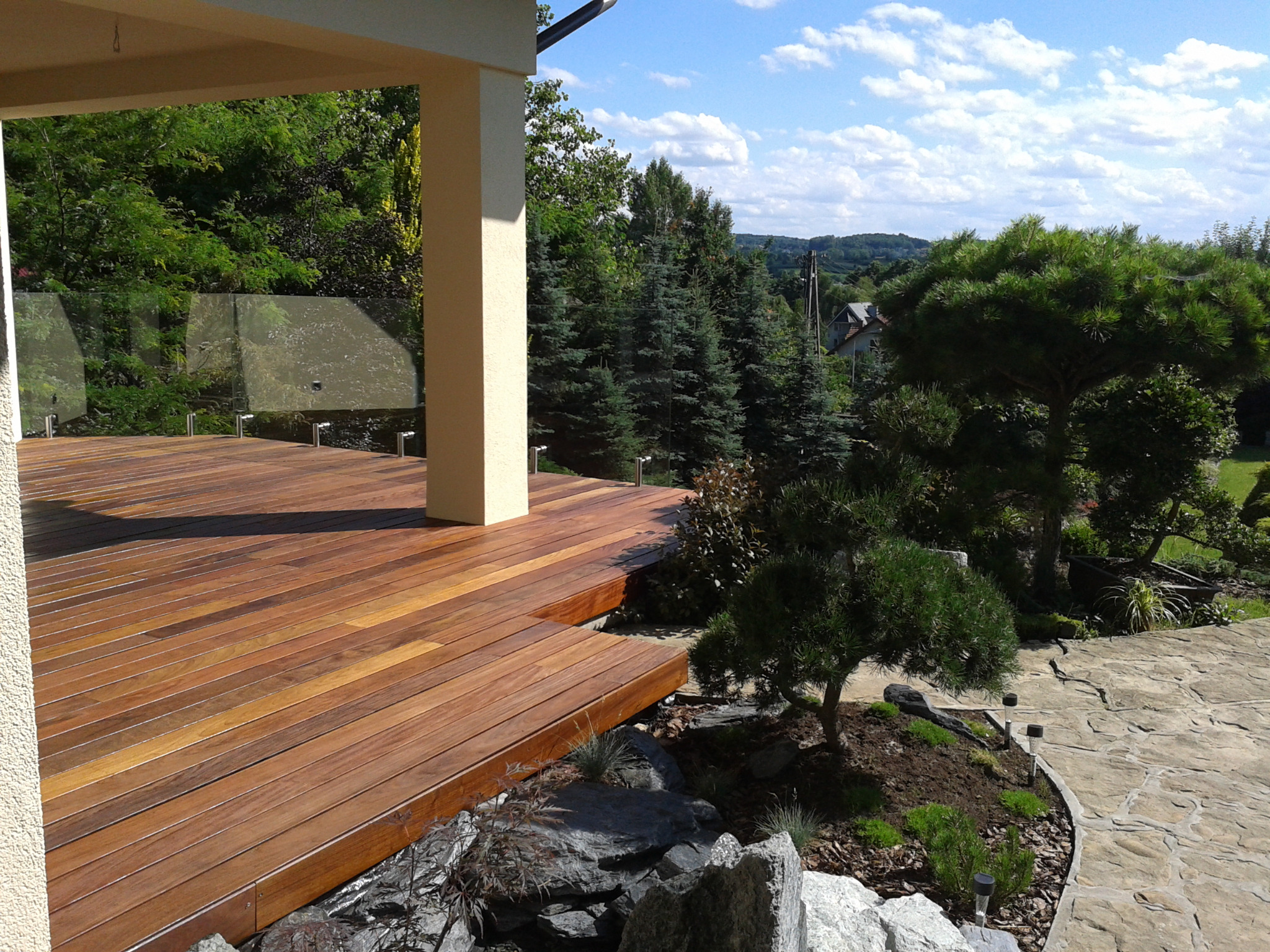 Nowoczesny taras z drewna egzotycznego, z widoczną szklaną balustradą i kolumną podtrzymującą zadaszenie, otoczony zielenią ogrodu skalnego i ścieżką z kamienia naturalnego.