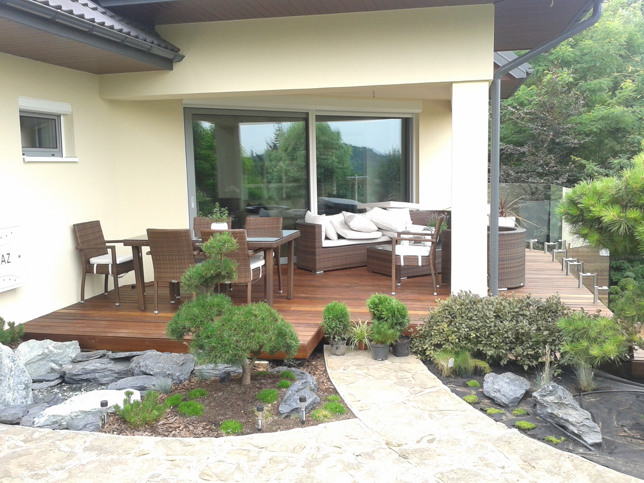 Zadaszony taras drewniany z meblami rattanowymi, ozdobnym skalniakiem z kamieniami i roślinami oraz ścieżką z kamienia prowadzącą do ogrodu.