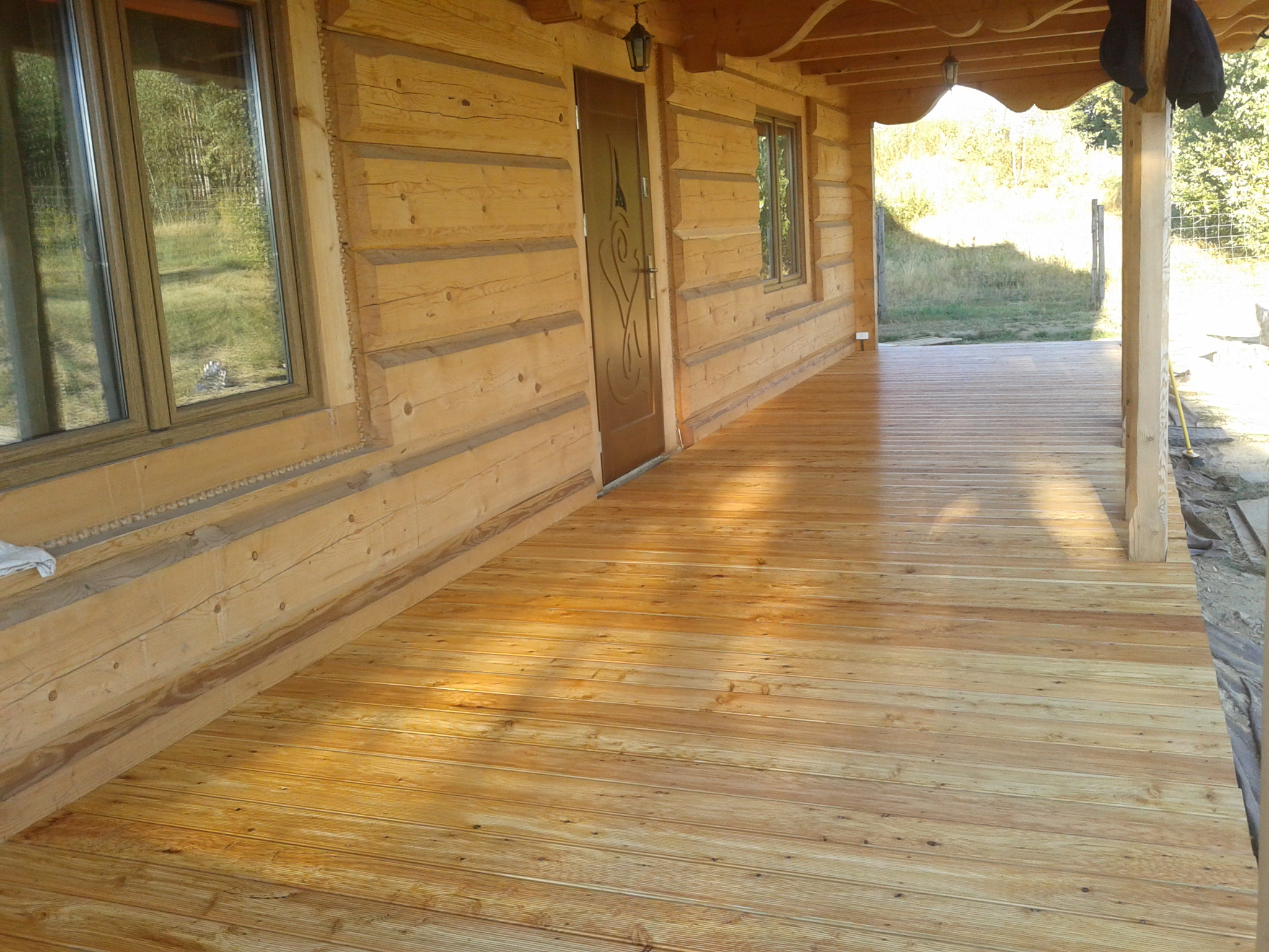 Zadaszony taras z desek drewnianych przy domu z bali, widoczne okna, drzwi z ozdobnym motywem i lampa na elewacji, w tle zieleń.