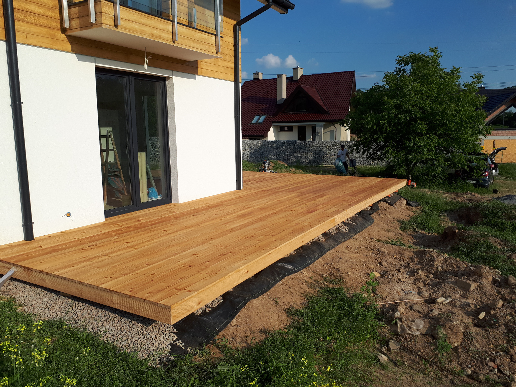 Nowo wybudowany taras z jasnego drewna przylegający do białego budynku z czarnymi oknami, widok na ogród i dom w tle.
