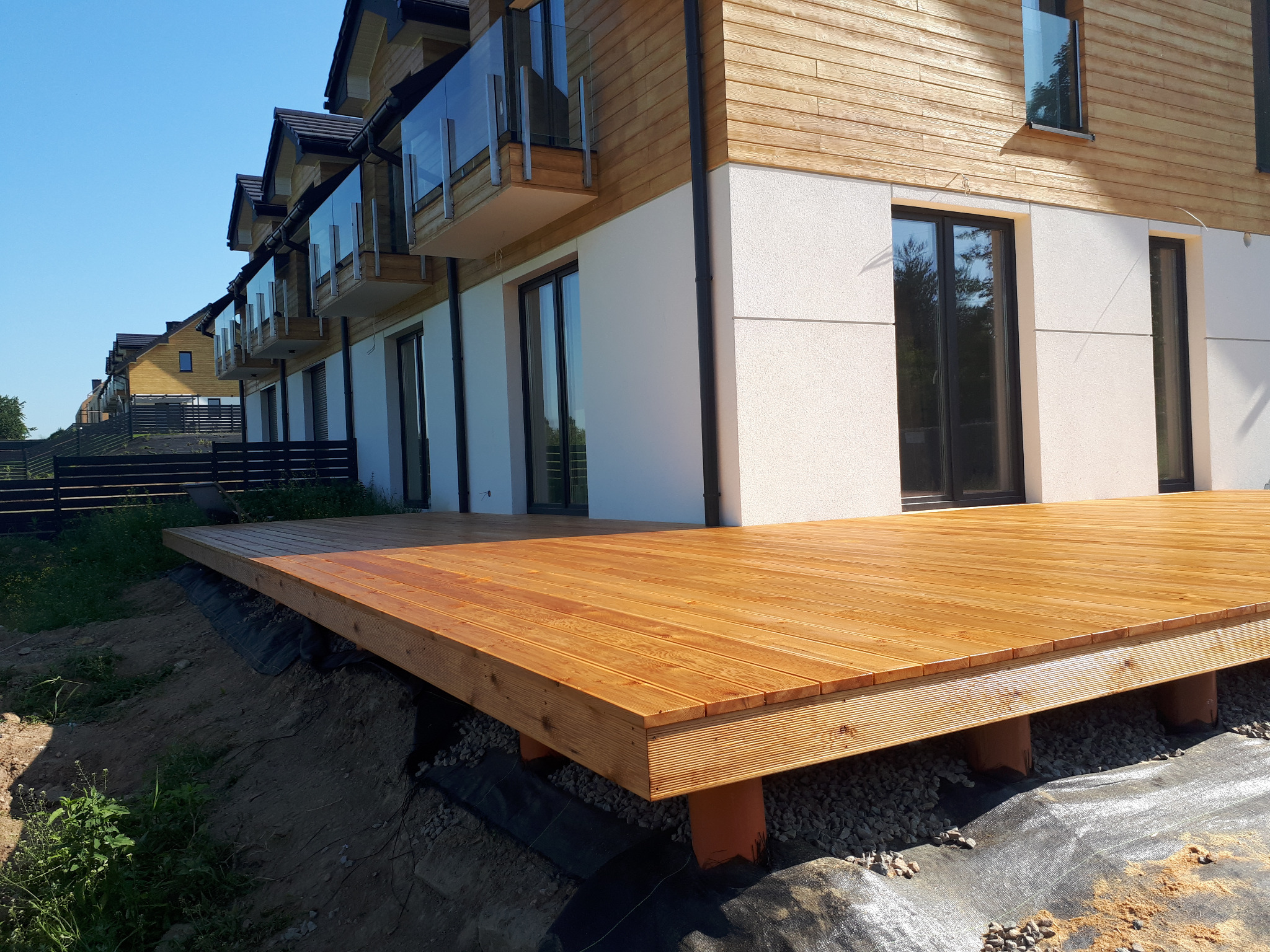 Nowoczesny taras z jasnego drewna przy szeregowcu, widoczne elementy konstrukcyjne i podłoże z kamienia.