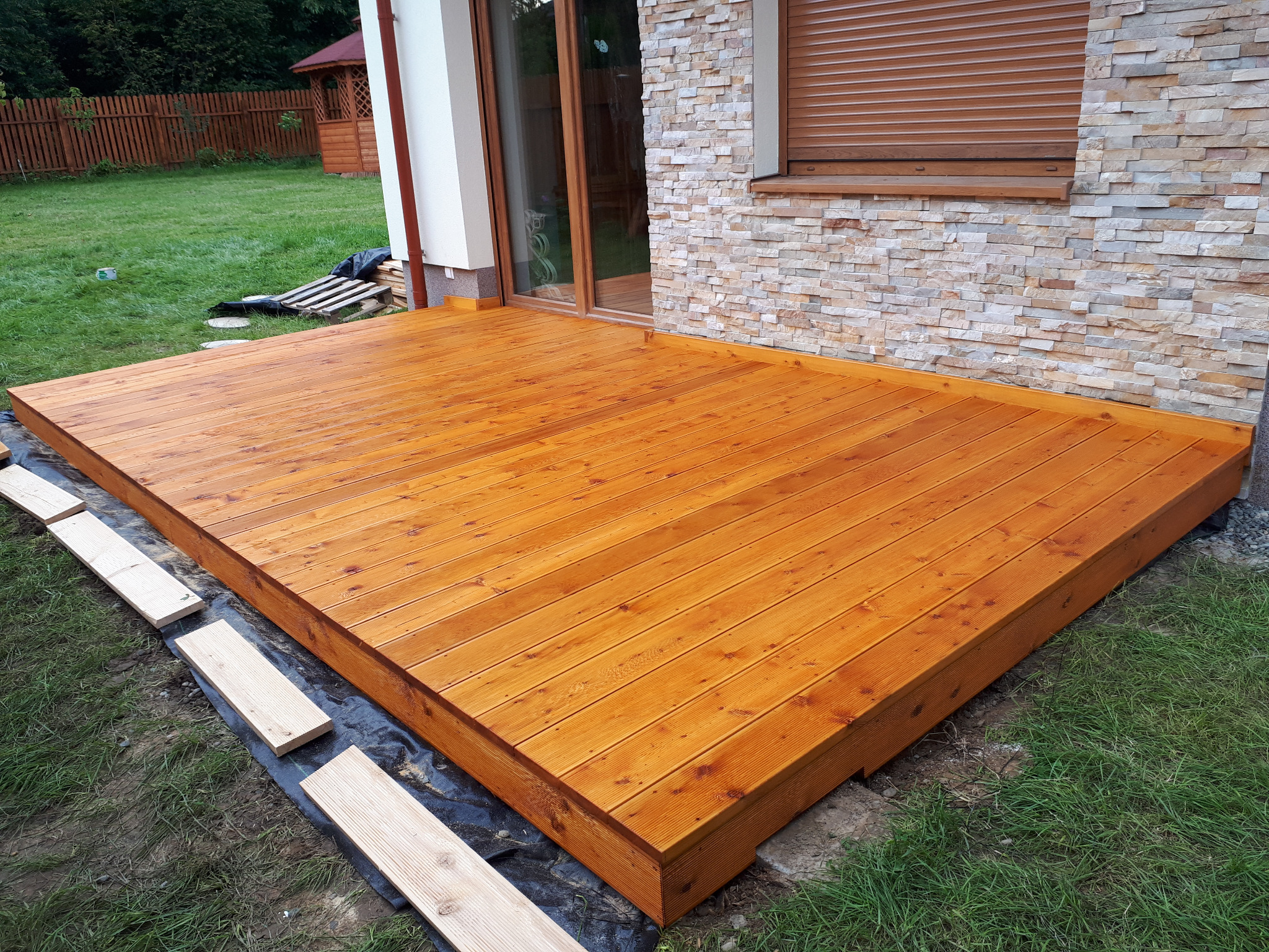 Nowo wybudowany taras z desek w ciepłym odcieniu brązu, przylegający do domu z elewacją z kamienia i brązowymi roletami, z widocznymi deskami przygotowanymi do dalszego montażu na czarnej folii.