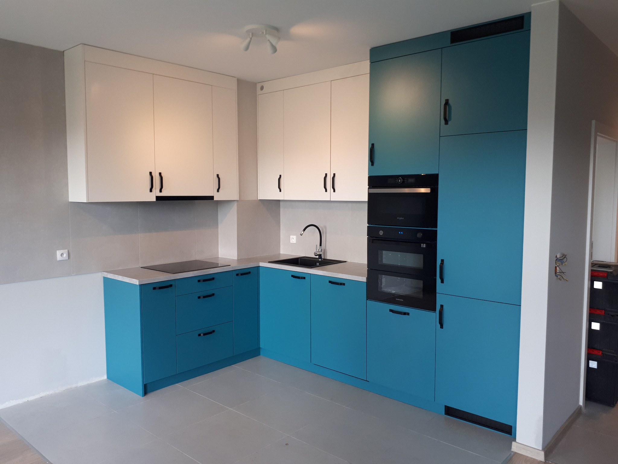 Nowoczesna kuchnia z białymi szafkami górnymi i turkusowymi dolnymi, czarne uchwyty, płyta indukcyjna i piekarnik w zabudowie, szara podłoga i ściany.