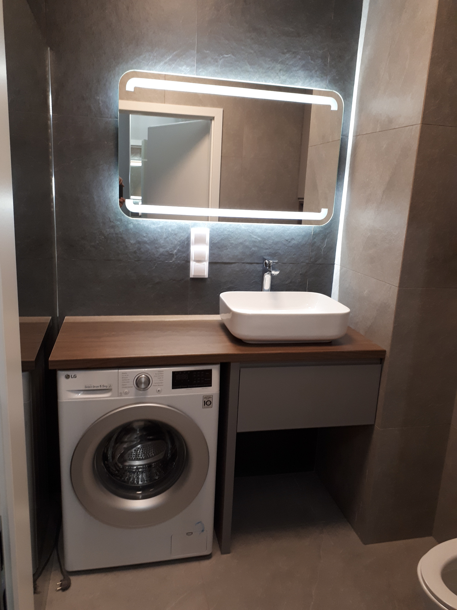 Nowoczesna łazienka z umywalką nablatową, podświetlanym lustrem i pralką w zabudowie pod blatem z ciemnego drewna.