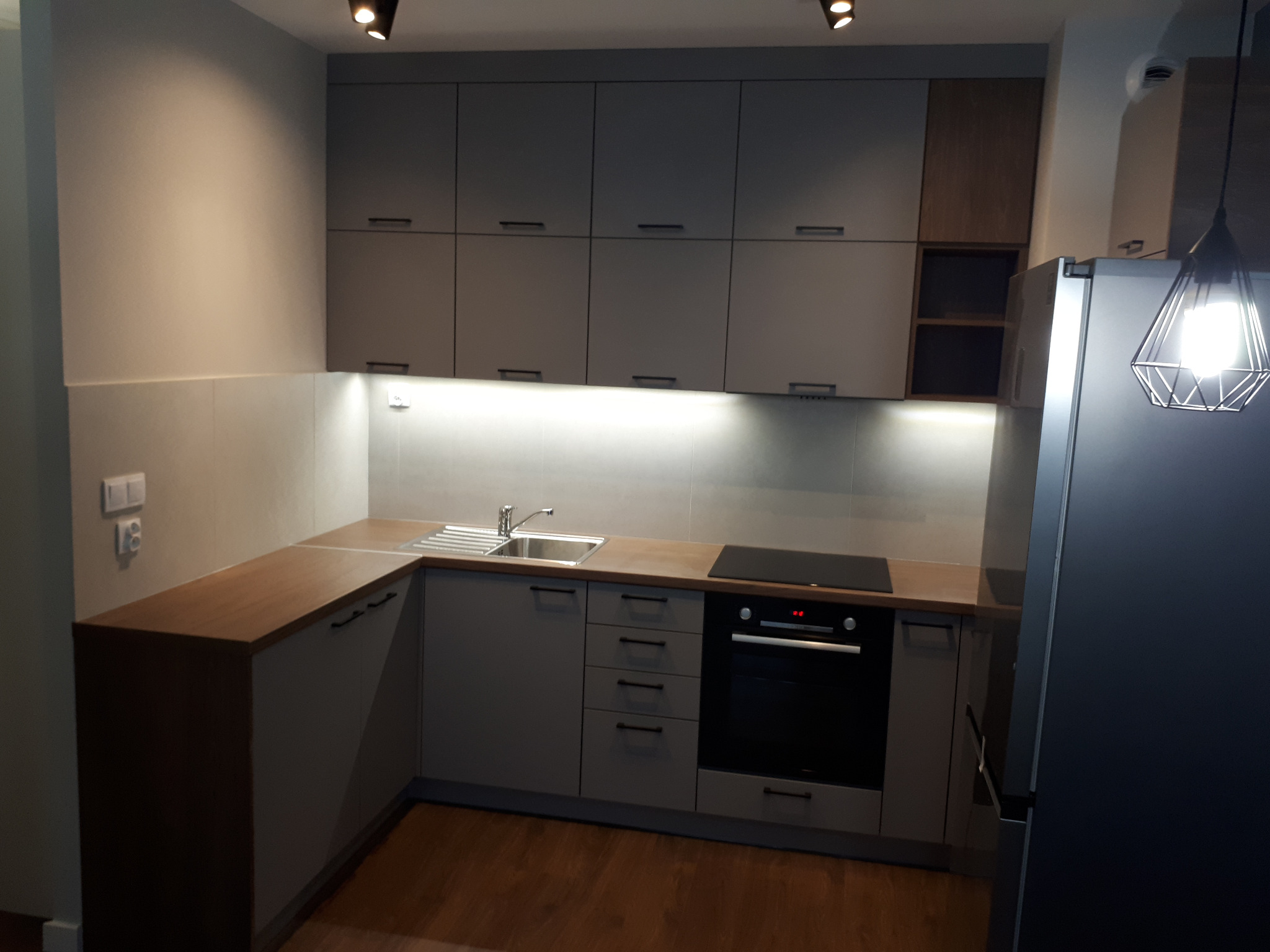 Nowoczesna kuchnia z szarymi szafkami, drewnianym blatem, oświetleniem LED pod szafkami wiszącymi i metalową lampą wiszącą, widziana z niskiego kąta.