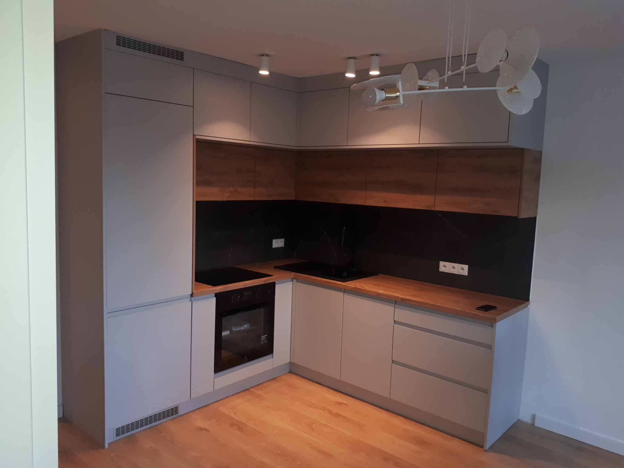 Nowoczesna kuchnia w kształcie litery L z szarymi frontami i drewnianymi blatami, czarny panel nad blatem, oświetlenie punktowe i designerski żyrandol, widok z niskiego kąta.