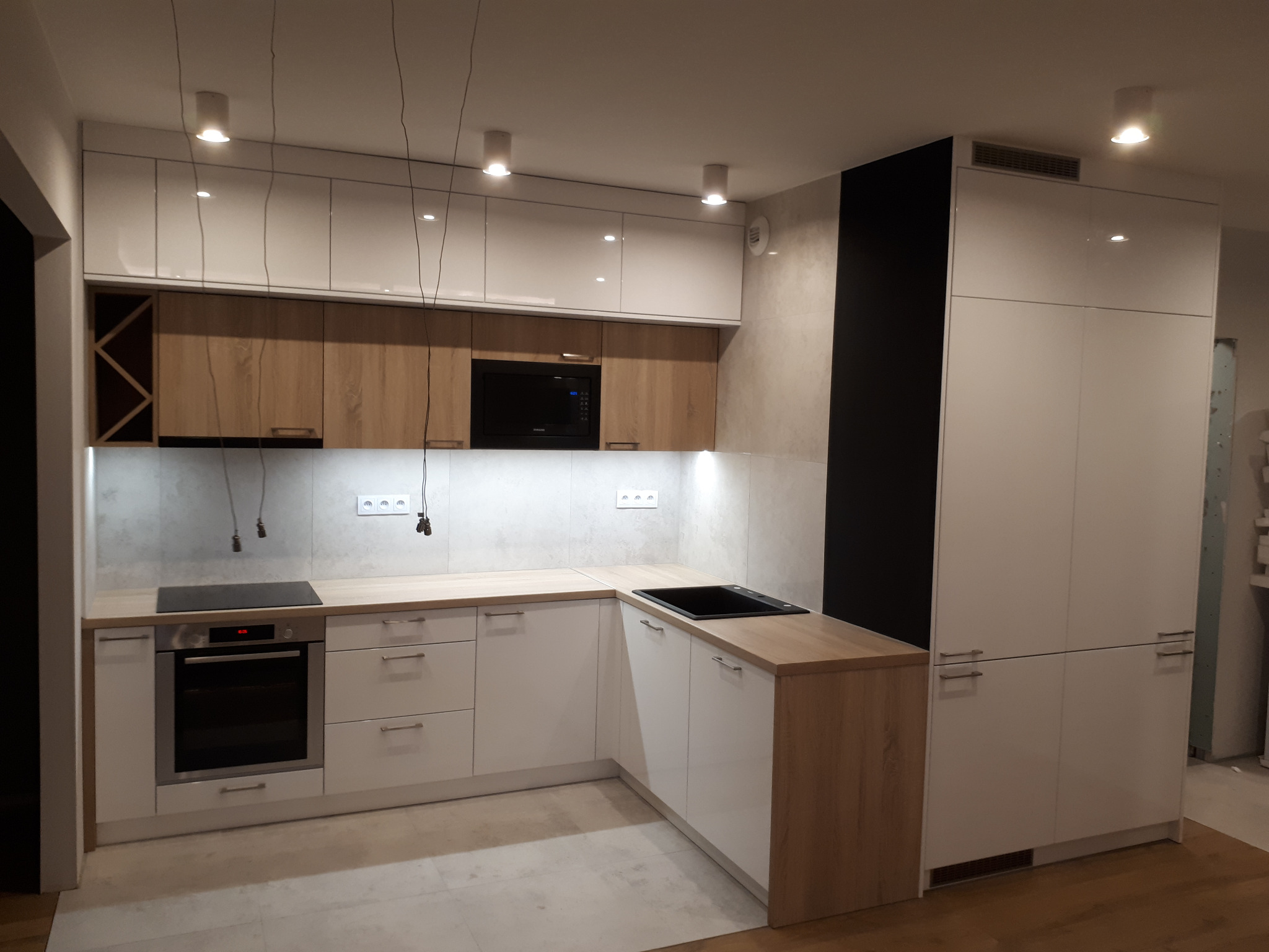 Nowoczesna kuchnia w odcieniach bieli i drewna, z jasnymi frontami szafek górnych i dolnych, drewnianymi blatami i czarnymi akcentami w postaci piekarnika, płyty indukcyjnej i wnęki na wino...