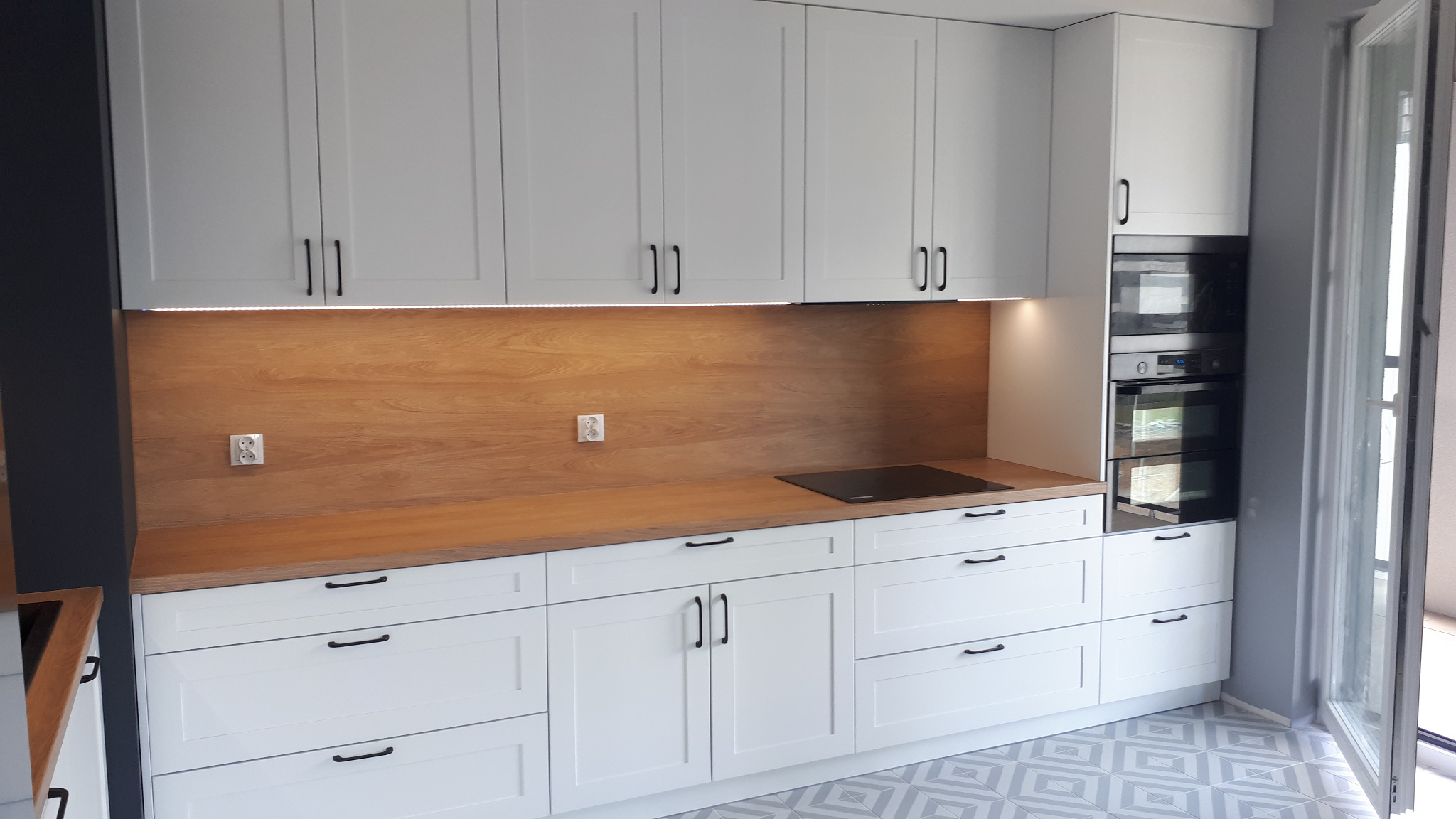 Nowoczesna kuchnia z białymi szafkami i drewnianym blatem, czarne uchwyty, płyta indukcyjna, piekarnik w zabudowie, gniazdka elektryczne na ścianie, podświetlenie LED pod szafkami górnymi, podłoga...
