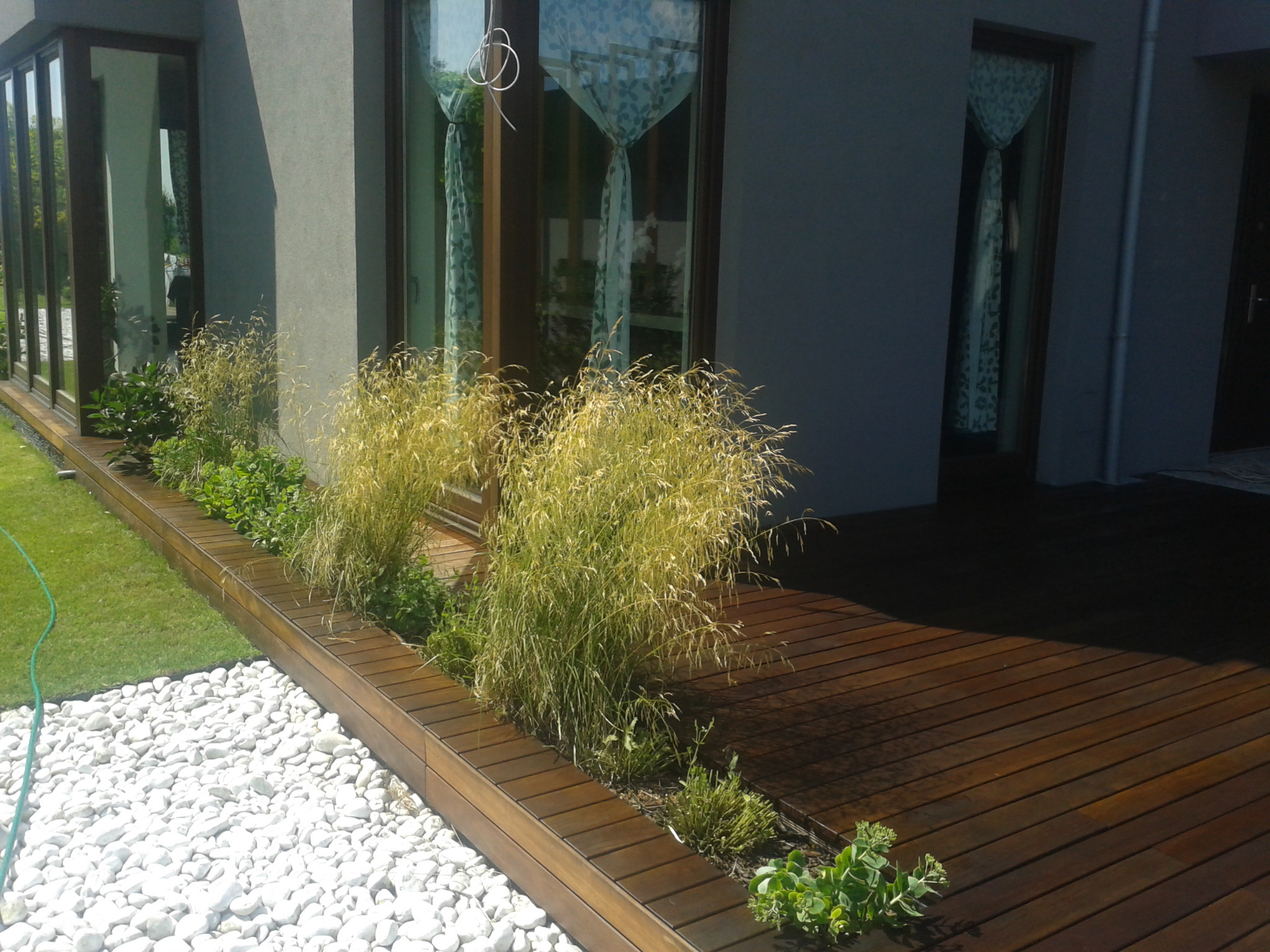 Ciemnobrązowy taras z desek, oddzielony od trawnika pasem białych kamieni i rabatą z zielenią ozdobną, przylegający do nowoczesnego domu z dużymi oknami.