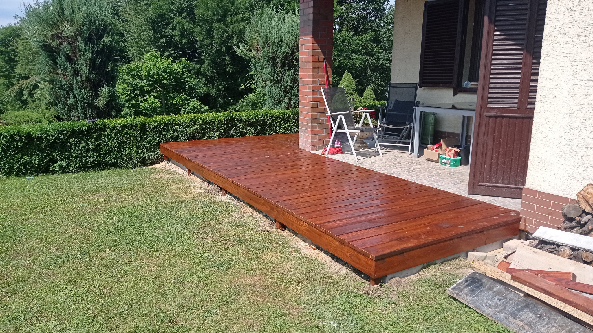 Świeżo polakierowany taras z drewna, przylegający do domu z ceglaną kolumną, otoczony trawnikiem i żywopłotem.