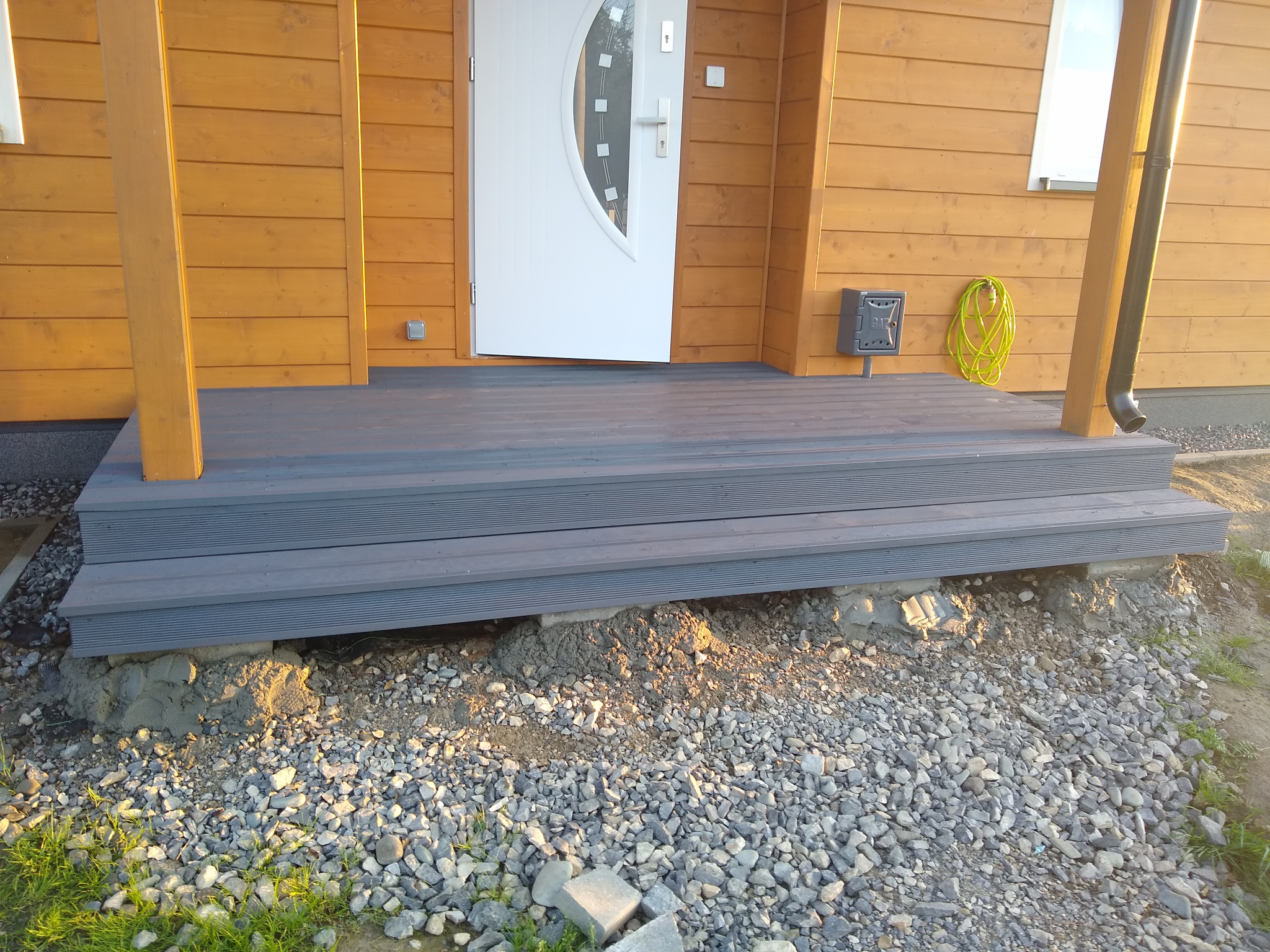 Schody wejściowe z kompozytu w kolorze szarym, prowadzące do domu z drewnianą elewacją, widoczne fundamenty i żwirowe podłoże.