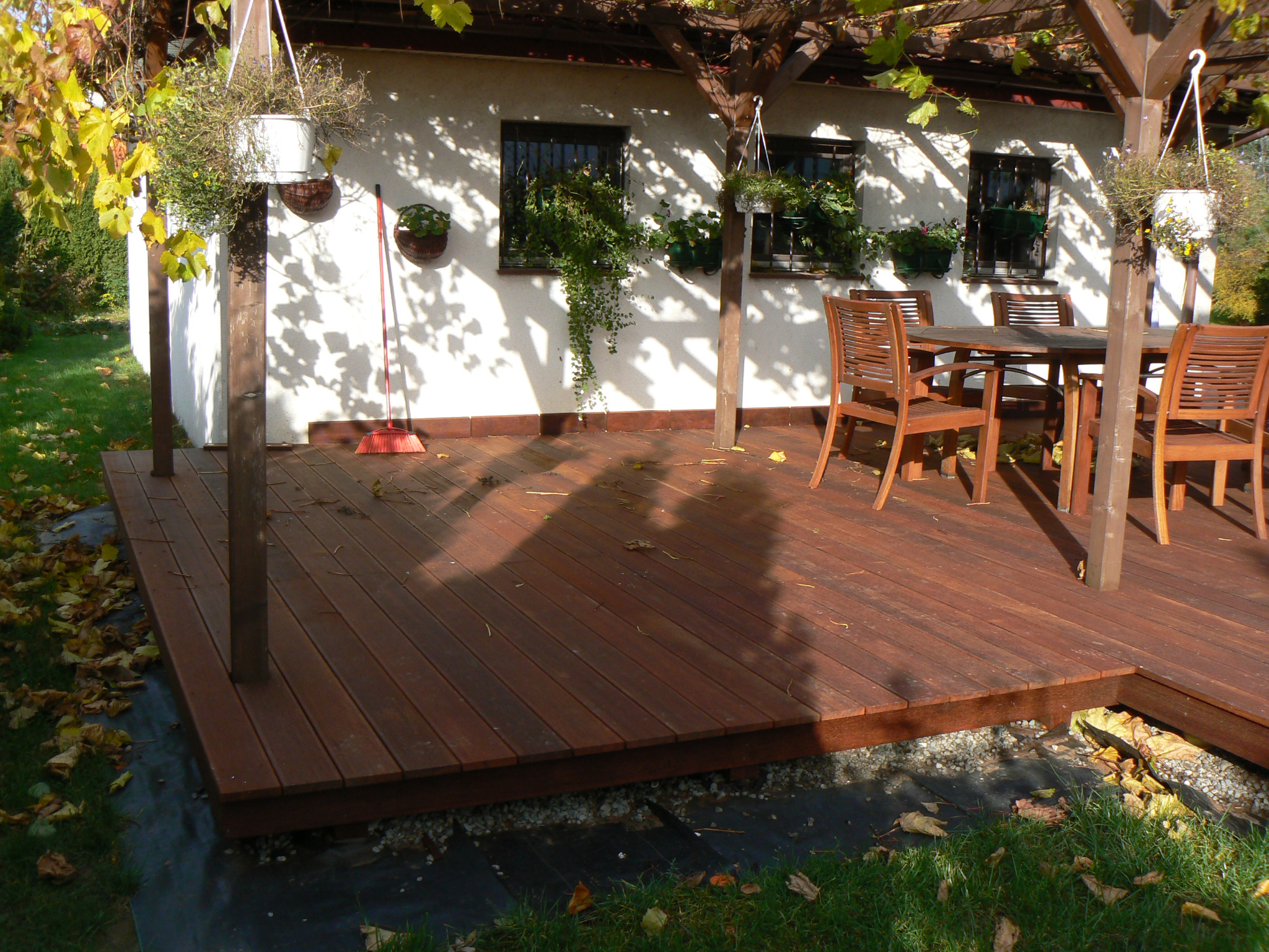 Zadaszony taras z drewna z brązowymi deskami, drewniane meble ogrodowe i wiszące donice z kwiatami, cień rzucany przez pergolę.