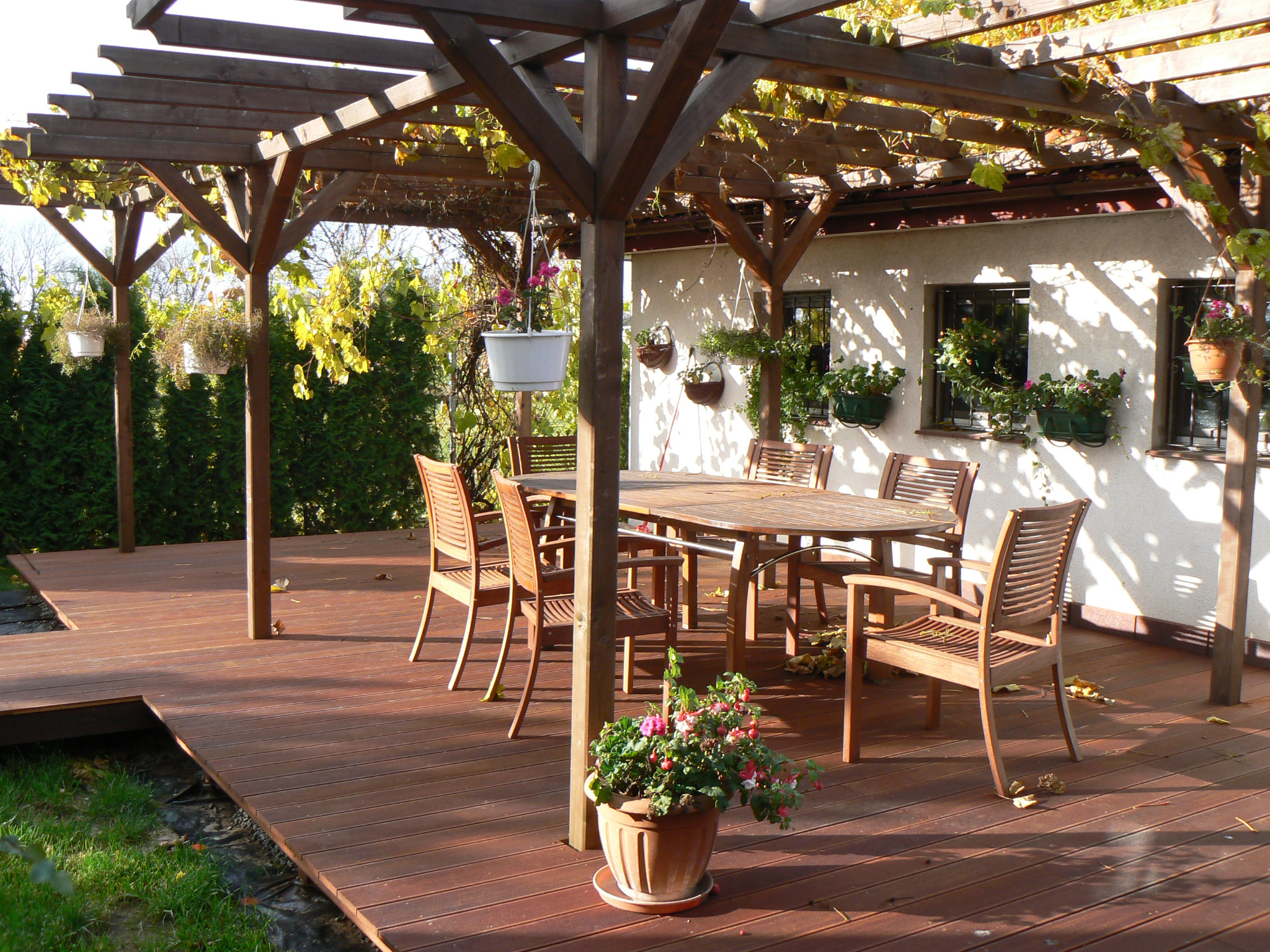Drewniany taras z pergolą, stół i krzesła ogrodowe, donice z kwiatami wiszące i stojące, jesienne liście na deskach, jasne światło słoneczne.