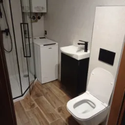 Nowoczesna łazienka z kabiną prysznicową typu walk-in, pralką, wiszącą toaletą i umywalką na szafce w kolorze czarnym, wykończona płytkami w odcieniach szarości i brązu.