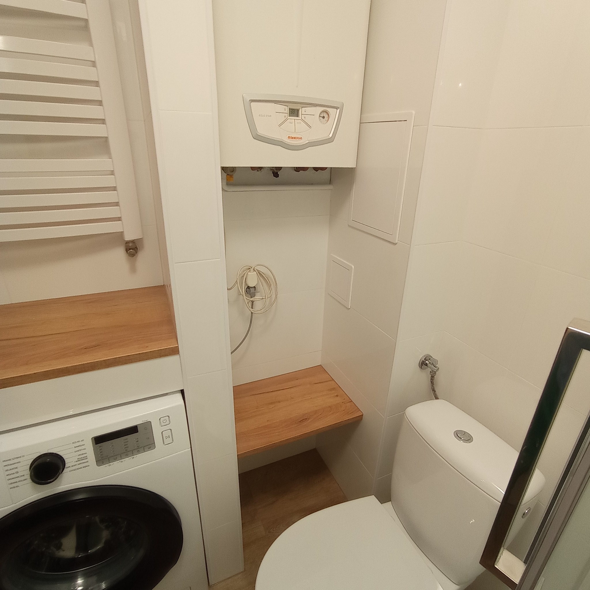 Kompaktowa łazienka z białą armaturą, pralką pod drewnianym blatem i wiszącym piecem gazowym Immergas Solo Star na ścianie z białych płytek.