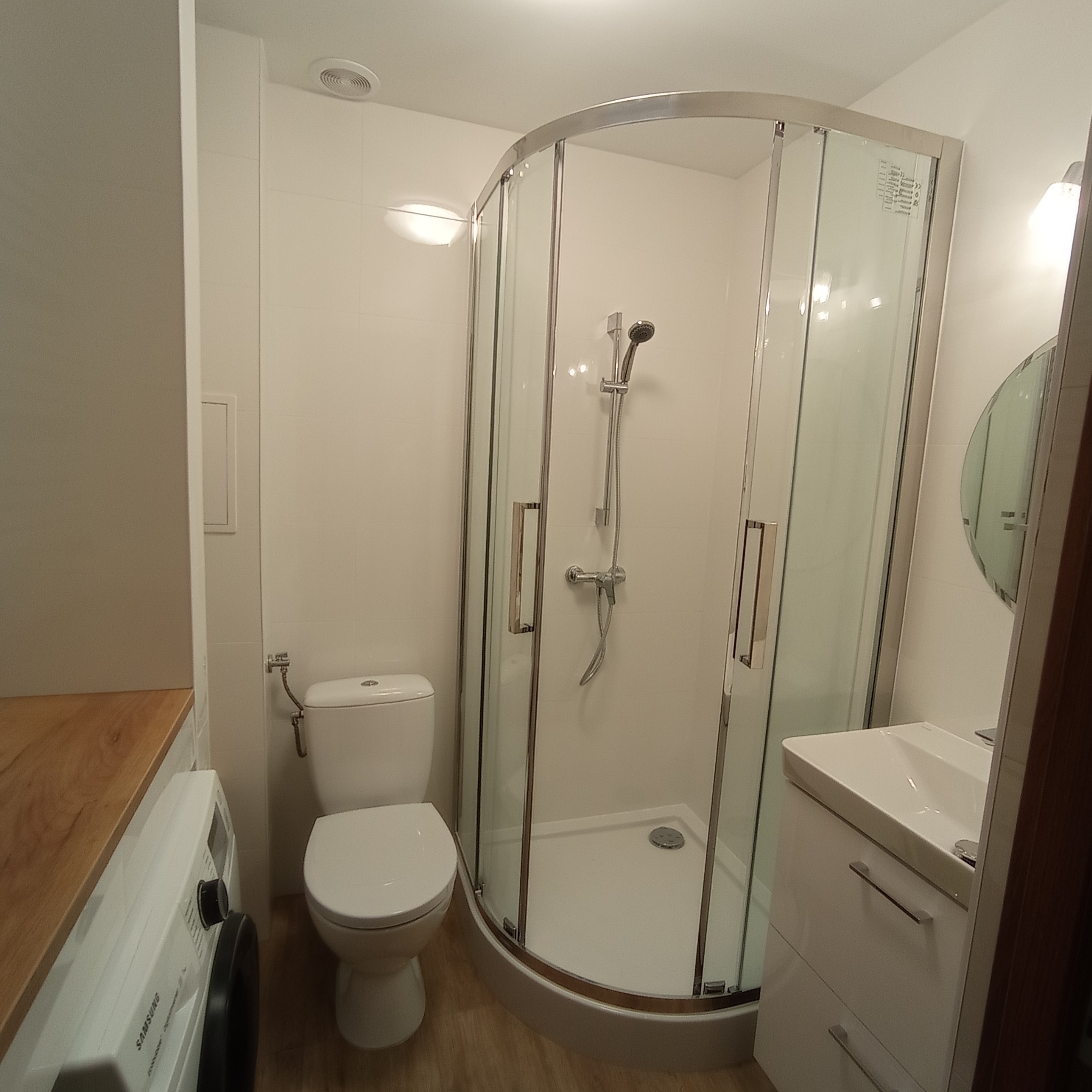 Nowoczesna łazienka z kabiną prysznicową, umywalką z szafką, toaletą i pralką pod drewnianym blatem, wykończona białymi płytkami.