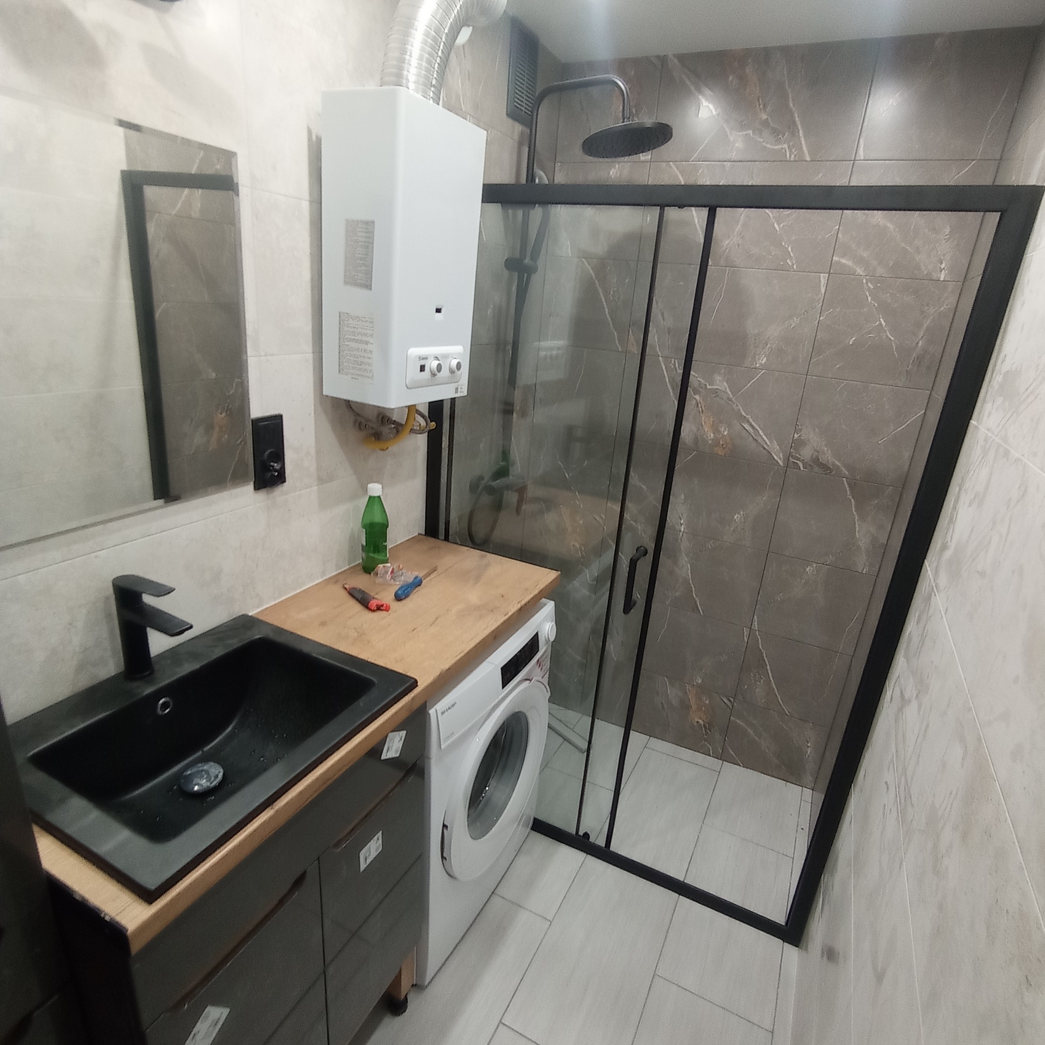 Nowoczesna łazienka z kabiną prysznicową z czarnymi ramami, umywalką wpuszczaną w blat, pralką oraz piecykiem gazowym, płytki imitujące marmur na ścianach.