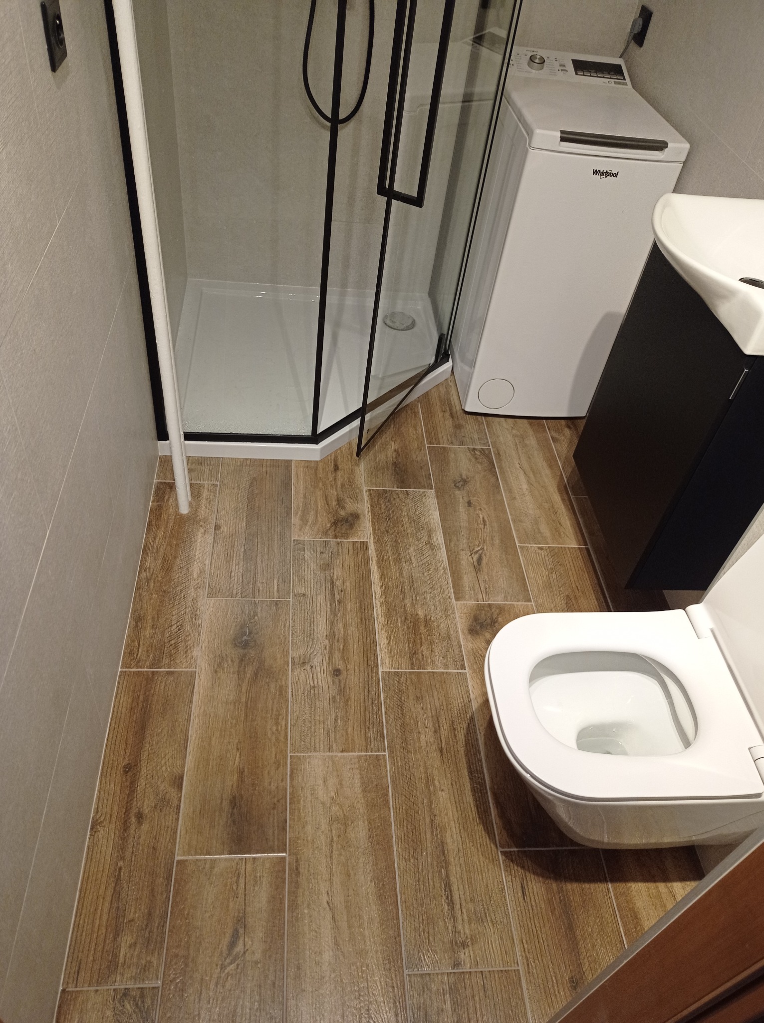 Wykończona łazienka z kabiną prysznicową z czarnymi profilami, pralką Whirlpool, umywalką z szafką i toaletą; podłoga wyłożona płytkami imitującymi drewno.
