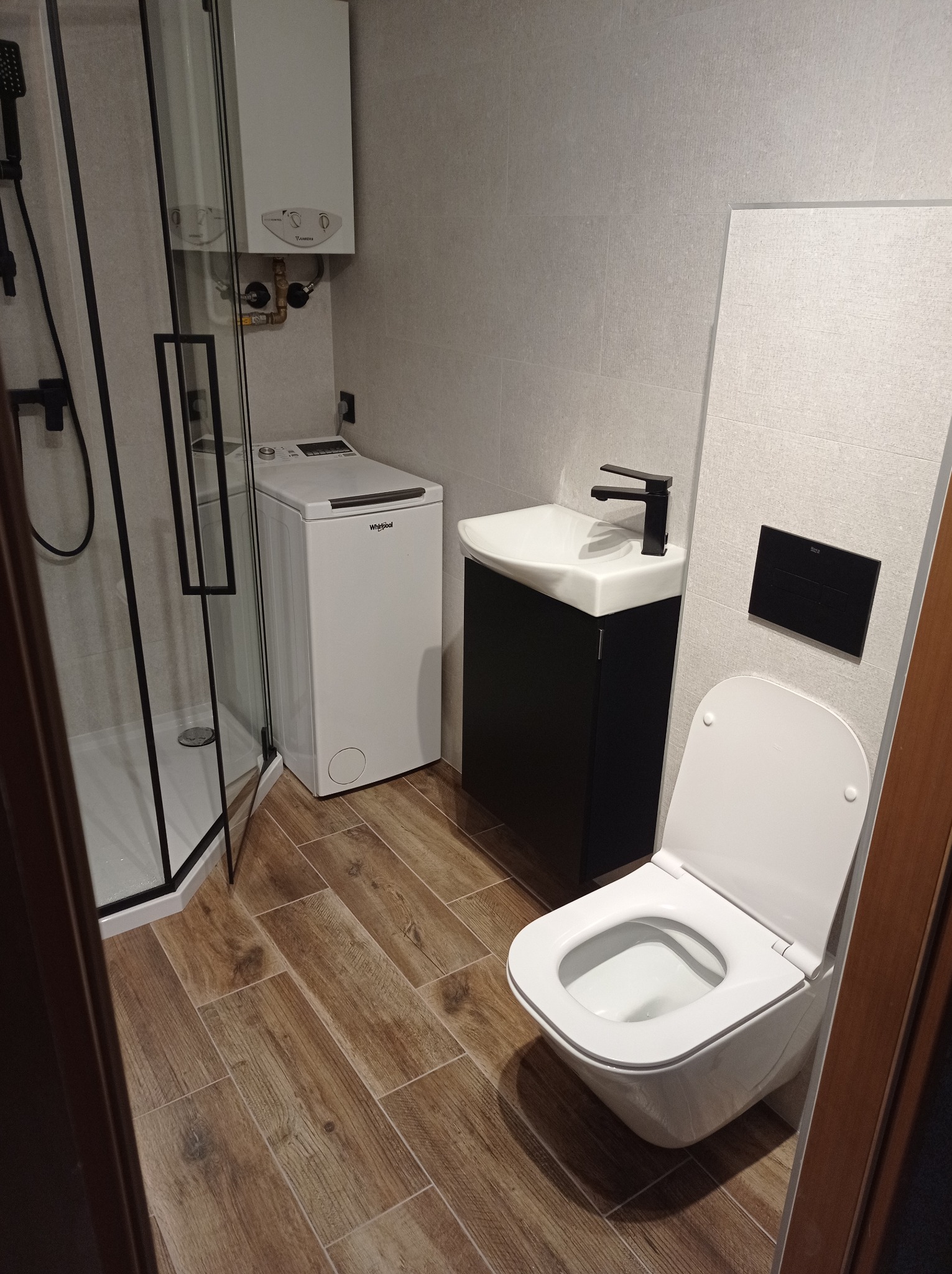 Nowoczesna łazienka z kabiną prysznicową typu walk-in, pralką, wiszącą toaletą i umywalką na szafce w kolorze czarnym, wykończona płytkami w odcieniach szarości i brązu.