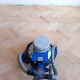 Parkiety Podłogi Układanie Cyklinowanie Renowacja Paweł Zdancewicz - Widok z góry na maszynę do obróbki parkietu na tle jasnej, ułożonej w jodełkę podłogi parkietowej, z widocznymi gniazdkami elektrycznymi na ścianie w tle.