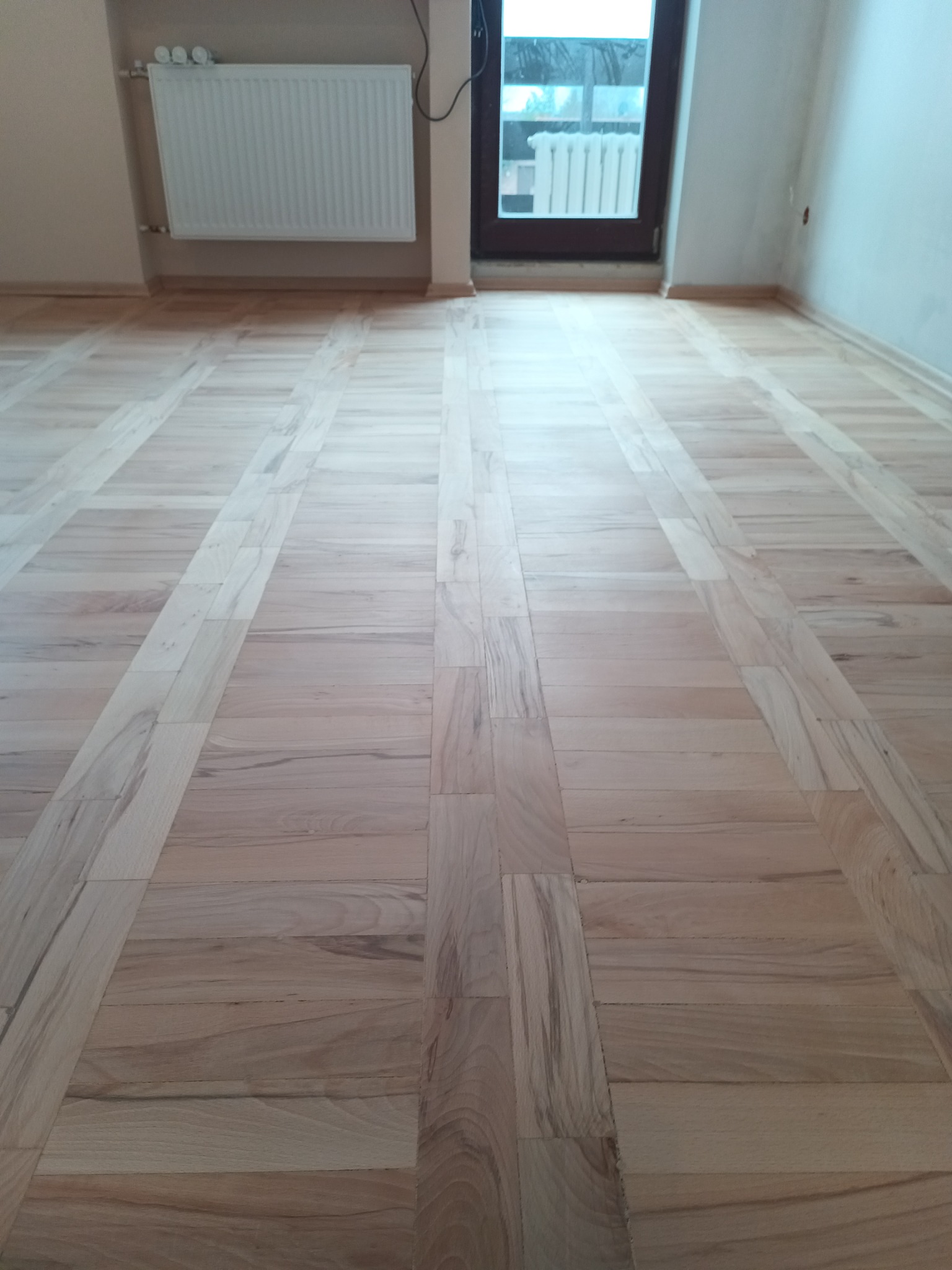 Jasny, ułożony parkiet w jodełkę francuską z widocznym grzejnikiem i oknem w tle.