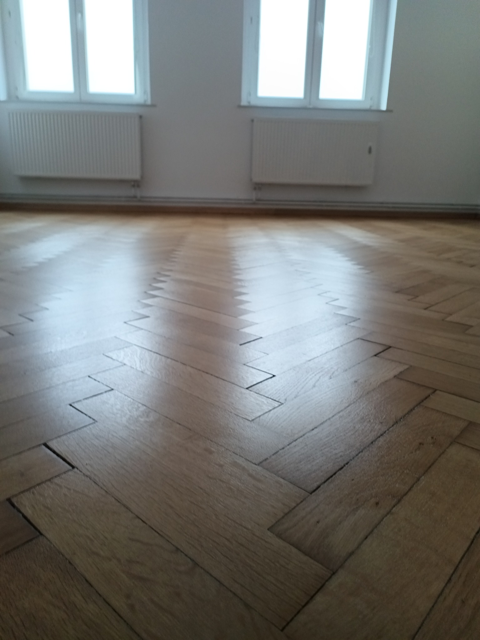Parkiet ułożony w jodełkę, po renowacji, z widocznymi promieniami światła odbijającymi się od lakierowanej powierzchni, w tle białe ściany i grzejniki.