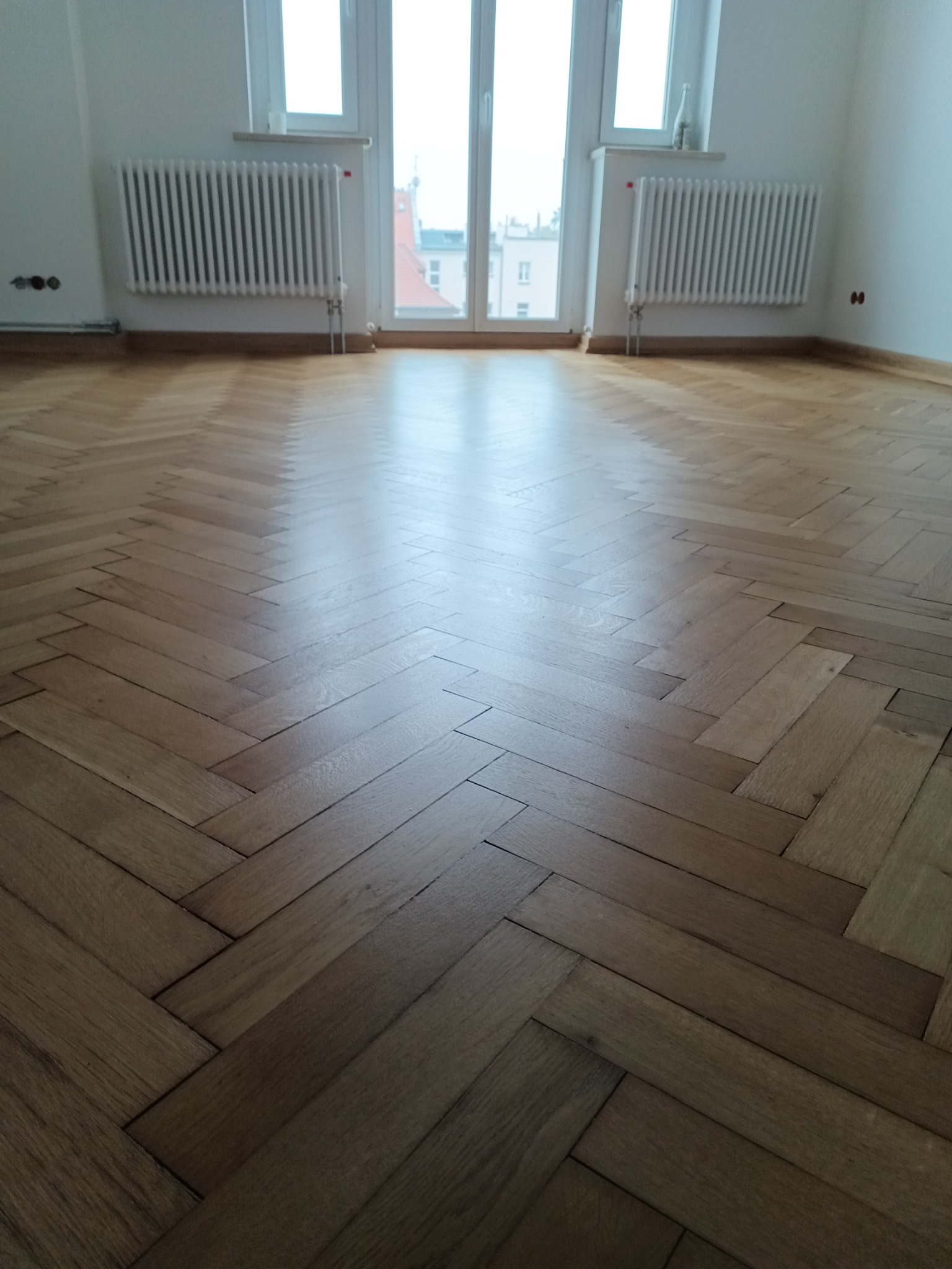 Parkiet ułożony w jodełkę w pustym pokoju z białymi ścianami, grzejnikami i oknem w tle.