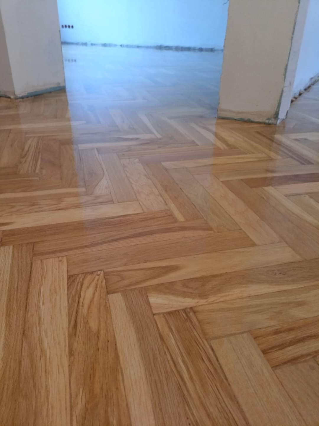 Błyszczący parkiet ułożony w jodełkę, świeżo po lakierowaniu, widoczny fragment ściany z niedoskonałościami.