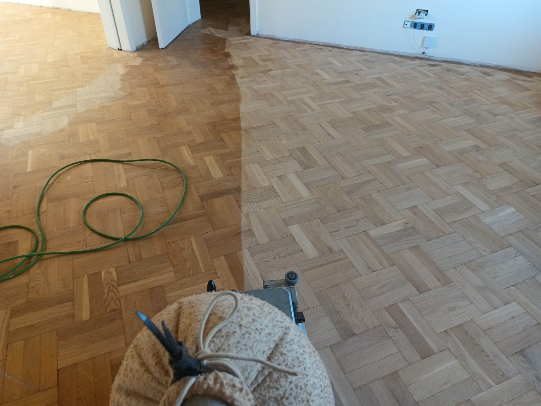 Parkiet ułożony w jodełkę, częściowo po procesie szlifowania, widoczna maszyna do cyklinowania i zielony przewód.