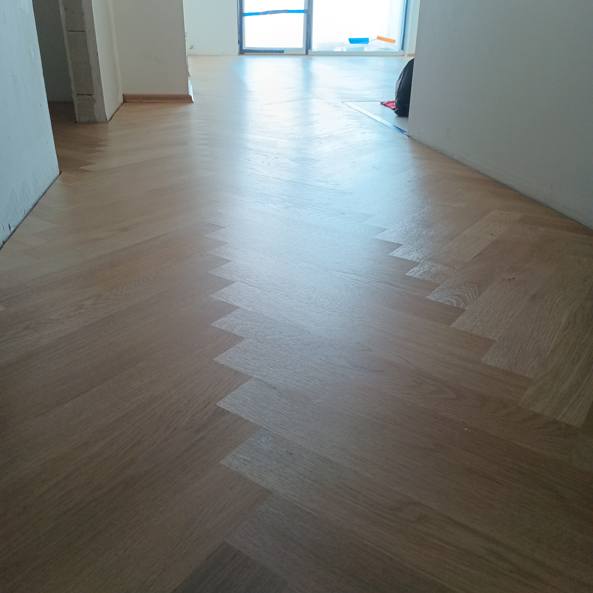 Jasny, parkiet ułożony w jodełkę w nowym mieszkaniu, widoczne przygotowane do malowania ściany i okna zabezpieczone folią.