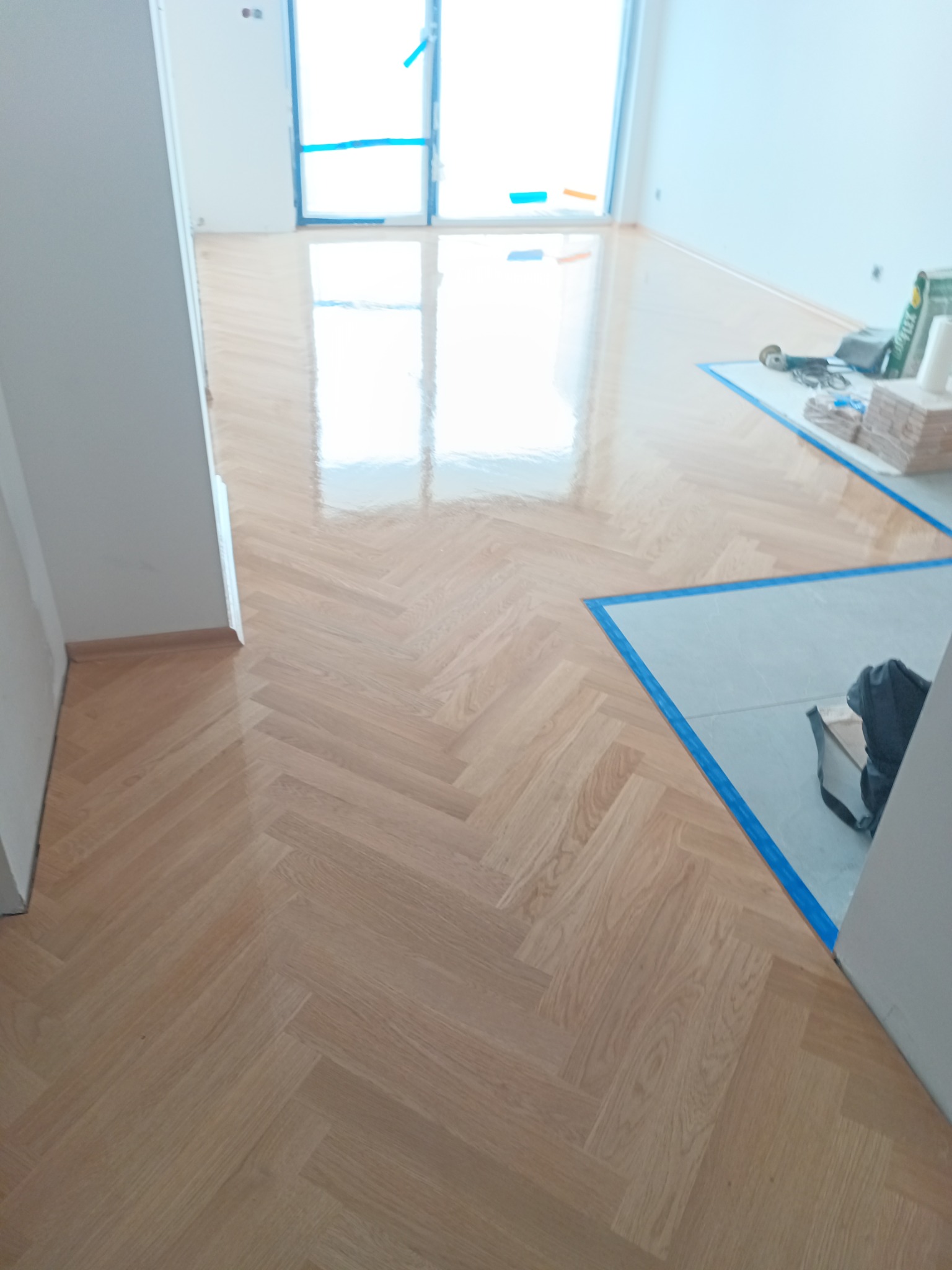 Parkiet ułożony w jodełkę, zabezpieczony niebieską taśmą malarską, widoczne materiały i narzędzia na podłodze w tle, jasne wnętrze.