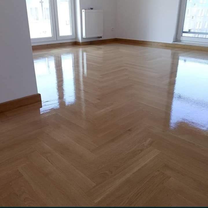 Błyszcząca, świeżo polakierowana podłoga parkietowa ułożona w jodełkę w pustym, jasnym pokoju z oknami.